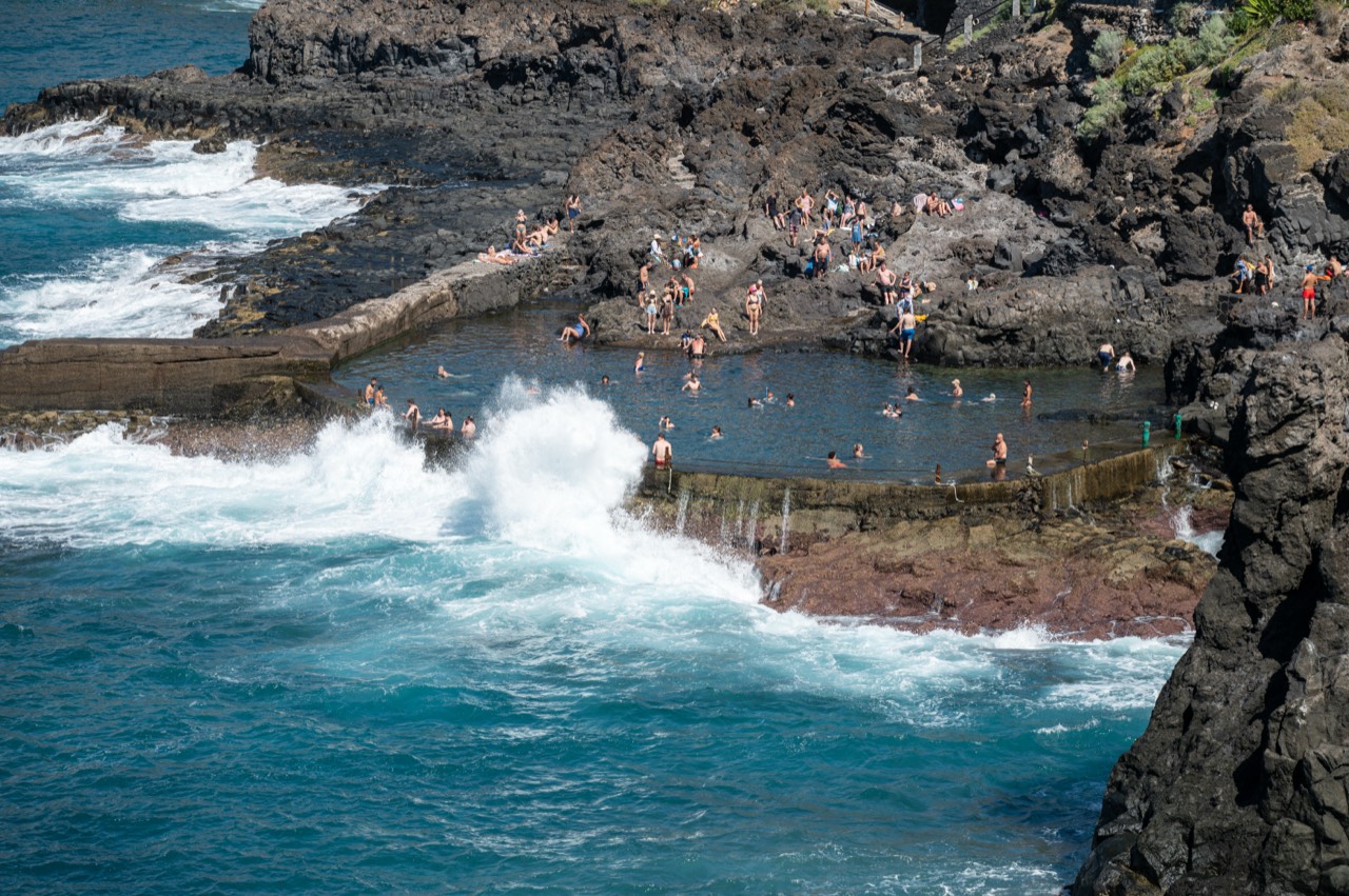 Bañistas en una piscina natural del litoral de Puerto de Santiago, con el oleaje rompiendo contra el muro protector